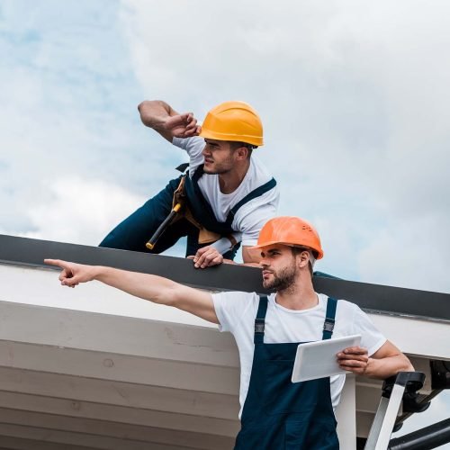 handsome bearded handyman in helmet holding digital tablet and pointing with finger near coworker