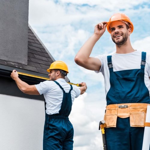 selective focus of happy repairman touching helmet and smiling near coworker